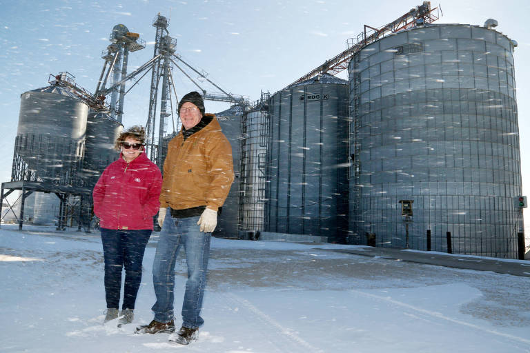 Um casal está posando em frente a grandes silos metálicos em um dia de neve. A mulher usa um casaco rosa e óculos escuros， enquanto o homem veste um casaco marrom e um gorro preto. A neve está caindo e o ambiente parece frio， com ventos visíveis na neve ao redor.