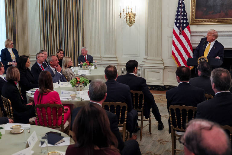 A imagem mostra uma reunião na Casa Branca， onde várias pessoas estão sentadas em torno de mesas com toalhas brancas. No fundo， há uma bandeira dos Estados Unidos e um candelabro. Um homem em pé， aparentemente falando， está à direita， enquanto os outros participantes escutam atentamente. A sala é decorada com cortinas e um grande quadro na parede.