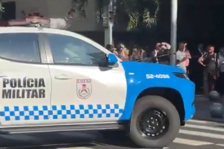 A imagem mostra uma viatura da Polícia Militar， com a parte frontal branca e a traseira azul， estacionada em uma faixa de pedestres. Ao fundo， há um grupo de pessoas observando， algumas delas segurando celulares. A viatura possui a inscrição 039;POLÍCIA MILITAR039; em destaque.