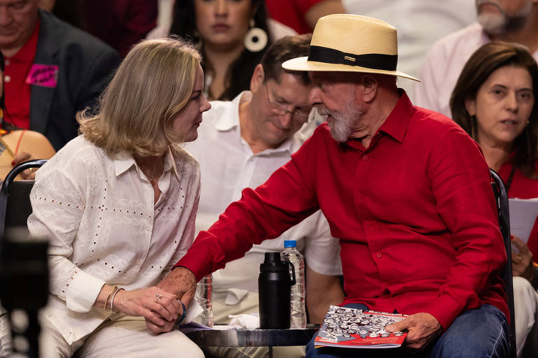 A imagem mostra duas pessoas sentadas em um evento， uma mulher com blusa branca e cabelo loiro， segurando a mão de um homem com camisa vermelha e chapéu. Ao fundo， outras pessoas estão assistindo ao evento， algumas com expressões atentas. O ambiente parece ser uma reunião ou comício político.