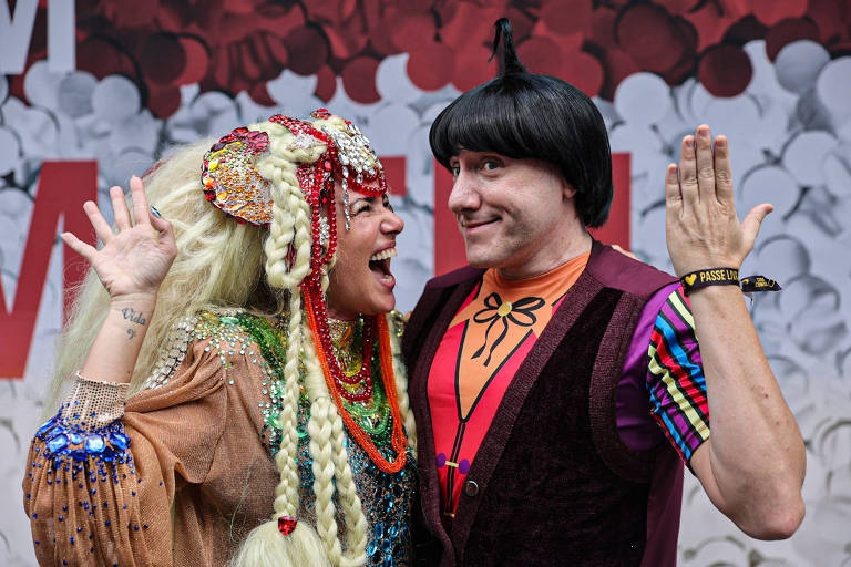 A imagem mostra duas pessoas vestidas com trajes coloridos e extravagantes. A mulher à esquerda tem cabelo longo e loiro， adornado com acessórios brilhantes， e usa um vestido decorado. O homem à direita tem cabelo escuro e usa uma camiseta colorida com um colete. Ambos estão sorrindo e fazendo gestos divertidos， com um fundo de papel picado em tons de vermelho e branco.