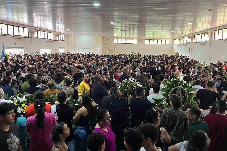 A imagem mostra uma grande multidão reunida em um espaço interno， possivelmente durante uma cerimônia ou velório. As pessoas estão em pé， algumas em grupos， e há arranjos de flores ao redor. O ambiente é bem iluminado， com janelas grandes ao fundo， e o teto é alto. A maioria das pessoas está vestida de maneira casual.