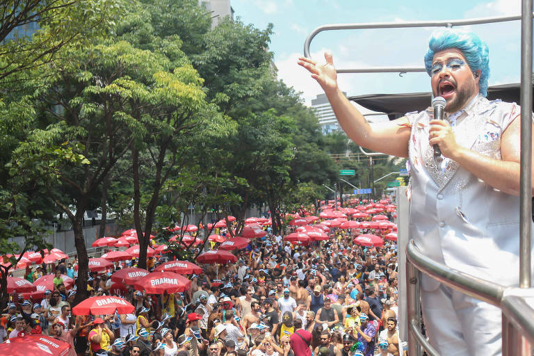 A imagem mostra uma grande multidão reunida em um evento de carnaval， com muitas pessoas usando roupas coloridas e algumas segurando guarda-sóis vermelhos. Em destaque， um homem com cabelo azul e roupas brilhantes está em um caminhão de som， falando para a multidão. Ao fundo， há árvores e prédios， indicando um ambiente urbano.