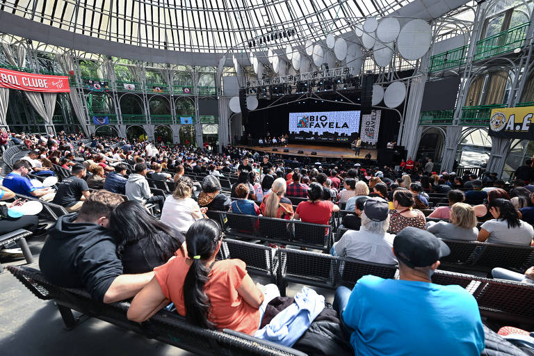 A imagem mostra um grande público reunido em um espaço coberto， com um palco ao fundo. O público está sentado em cadeiras， e a maioria das pessoas está voltada para o palco， onde há uma tela grande com o texto 039;BIO FAVELAS039;. O ambiente é iluminado e possui uma estrutura de vidro no teto， permitindo a entrada de luz natural.