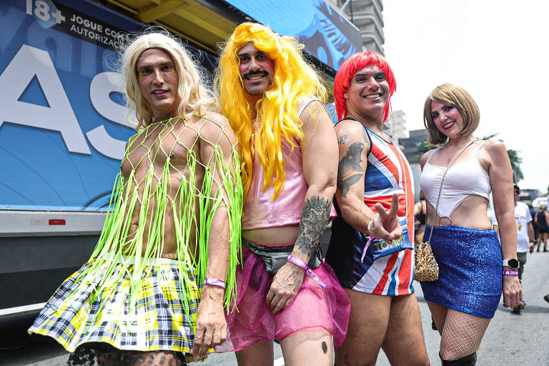 Quatro pessoas posando para a foto em um evento de rua, vestindo roupas coloridas e extravagantes. Dois homens estão usando perucas longas, um com cabelo loiro e outro com cabelo vermelho. Um deles está vestido com uma rede verde e uma saia rosa, enquanto o outro usa uma camiseta com a bandeira do Reino Unido e shorts. A mulher à direita está usando uma blusa branca e uma saia curta brilhante. Ao fundo, há um caminhão com a palavra 'AS' visível.