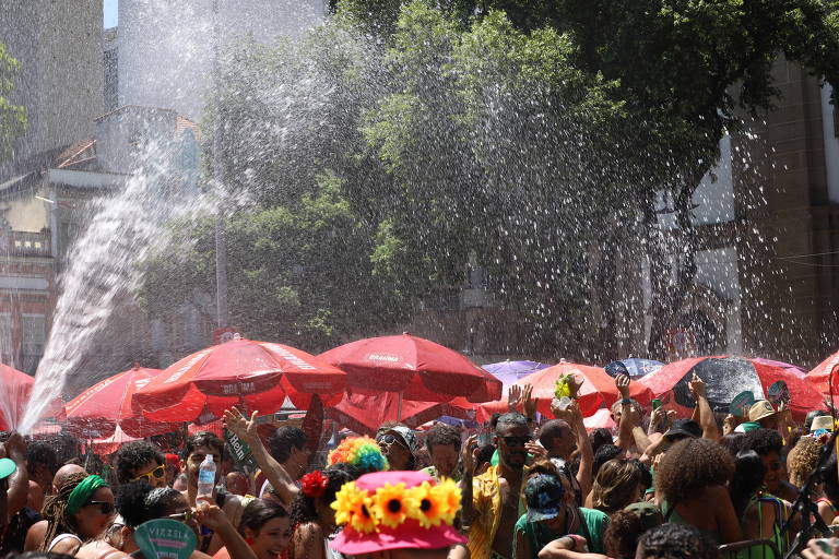 A imagem mostra uma grande multidão celebrando em um evento ao ar livre, possivelmente durante o Carnaval. As pessoas estão se divertindo sob um chuveiro de água, com algumas levantando os braços. Há várias sombrinhas vermelhas ao fundo, e a cena é iluminada pelo sol. Muitas pessoas usam roupas coloridas e acessórios festivos.