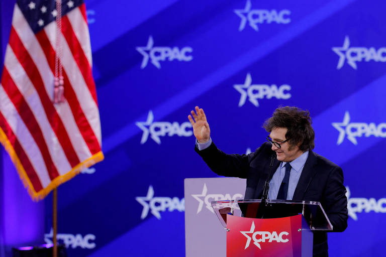 A imagem mostra um homem em um palco durante o evento CPAC， com uma bandeira dos Estados Unidos ao fundo. O homem está de pé， com um microfone à sua frente， acenando com a mão direita. O fundo é decorado com um padrão azul e o logotipo do CPAC em branco.