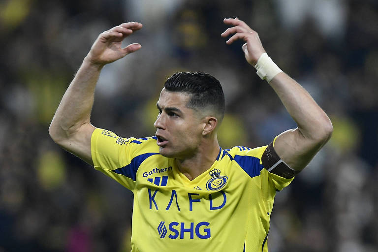 Um jogador de futebol está em campo， vestindo uma camisa amarela com detalhes azuis. Ele levanta as mãos， expressando uma reação， possivelmente de frustração ou celebração. O jogador usa uma faixa no braço e parece estar concentrado na partida. O fundo é desfocado， mas sugere um estádio com torcedores.