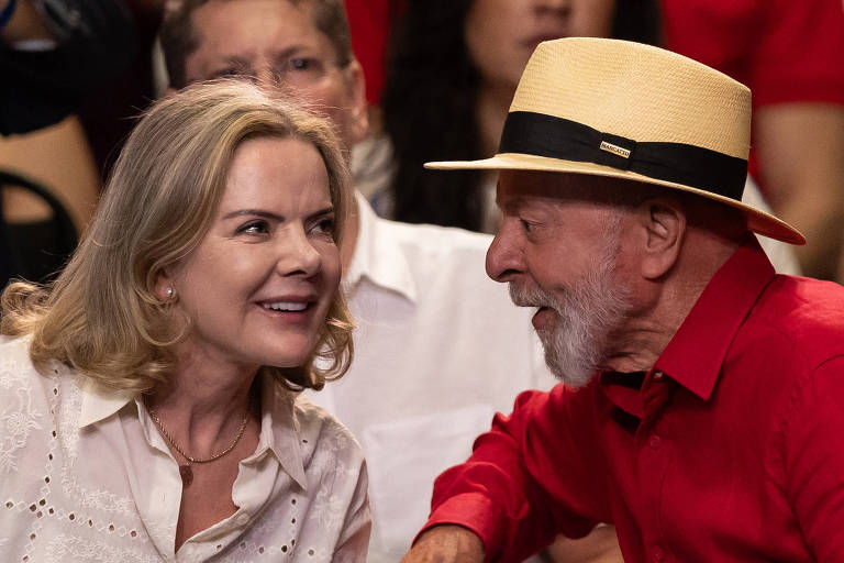 A imagem mostra duas pessoas conversando em um evento. À esquerda， uma mulher com cabelo loiro e ondulado， vestindo uma blusa clara com detalhes em renda. À direita， um homem idoso com barba branca， usando uma camisa vermelha e um chapéu de palha com uma faixa preta. Ao fundo， outras pessoas estão visíveis， algumas com roupas em tons de vermelho.