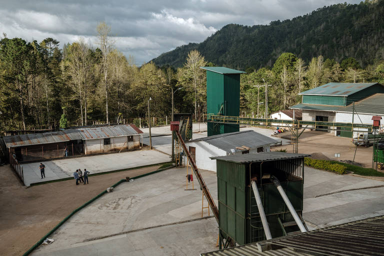 A imagem mostra uma área industrial cercada por vegetação densa e montanhas ao fundo. No centro, há estruturas metálicas, incluindo um grande silo verde e um sistema de transporte. Algumas pessoas estão visíveis em uma das áreas pavimentadas, enquanto outras estão em movimento. O céu está nublado, sugerindo um clima ameno.