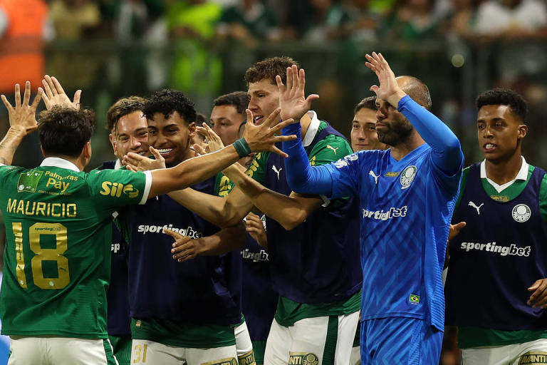 Um grupo de jogadores de futebol celebra um gol. Eles estão usando uniformes verdes e azuis， com alguns jogadores levantando as mãos em comemoração. O goleiro， vestido de azul， está no centro da imagem， cercado por seus companheiros. O fundo mostra uma torcida animada.