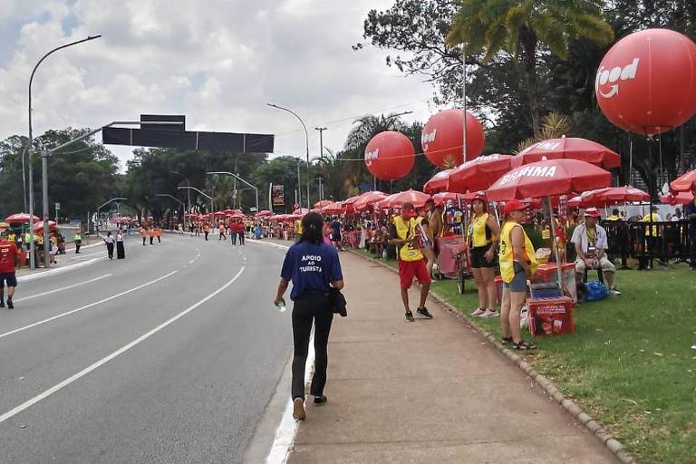 A imagem mostra uma rua com pessoas caminhando e barracas montadas ao longo da calçada. Há várias pessoas vestindo camisetas amarelas e algumas barracas com toldos vermelhos. Balões vermelhos estão visíveis， e ao fundo， há uma construção e árvores. O céu está parcialmente nublado.