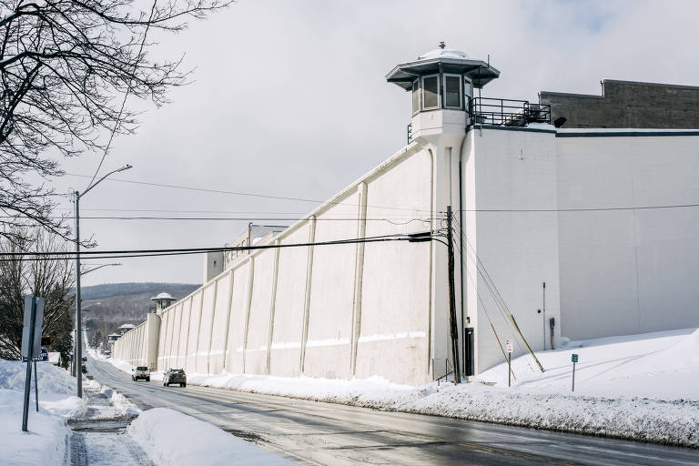 A imagem mostra uma prisão com uma alta parede branca e uma torre de vigilância no topo. A cena é em um dia nublado， com neve acumulada ao longo da estrada e nas laterais. Há um carro passando na estrada em frente à prisão， que é cercada por árvores sem folhas.