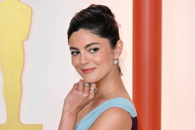 Uma mulher está posando em um evento de gala, vestindo um vestido longo com um corpete azul claro e uma saia roxa. Ela está com uma mão no queixo e a outra braço cruzado, olhando para a câmera. Ao fundo, há uma figura de estatueta dourada.