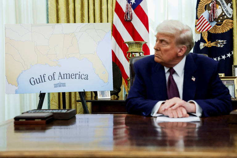 A imagem mostra um homem sentado à mesa em um ambiente formal， provavelmente na Sala Oval. Ele tem cabelo loiro e está usando um terno escuro com uma gravata. Ao fundo， há uma bandeira dos Estados Unidos e um brasão presidencial. À sua esquerda， há um quadro com um mapa que diz 039;Gulf of America039;. A mesa tem um objeto preto em sua superfície.