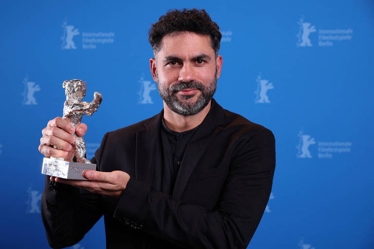 Um homem com barba e cabelo encaracolado， vestindo uma jaqueta preta， está segurando um troféu em forma de urso em frente a um fundo azul. O troféu é prateado e ele parece estar sorrindo.