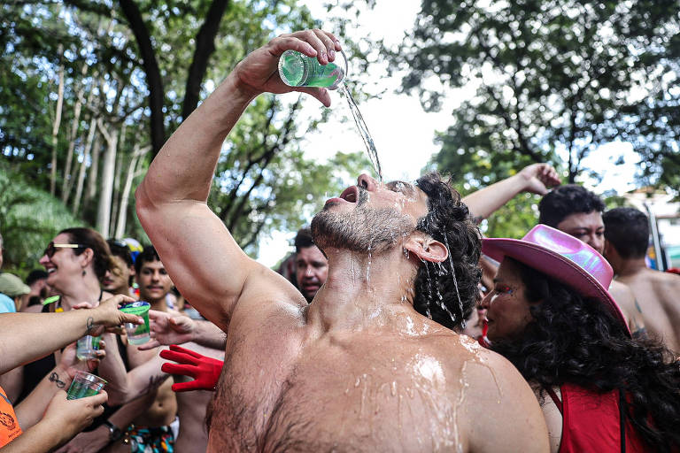 Um homem sem camisa está bebendo de uma lata， inclinando a cabeça para trás enquanto a bebida escorre pelo seu rosto. Ao redor dele， várias pessoas estão se divertindo， algumas segurando latas de bebida. O ambiente é festivo， com árvores ao fundo e pessoas sorrindo e dançando.