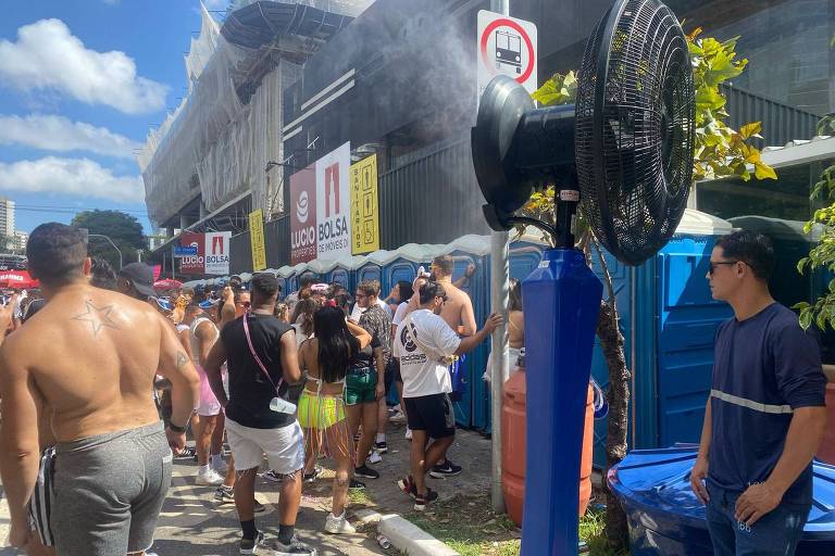 A imagem mostra um evento ao ar livre com várias pessoas em uma fila. Algumas pessoas estão usando roupas de verão， enquanto outras estão em trajes de banho. Há um ventilador grande ao lado da fila， que parece estar borrifando água. Ao fundo， há uma construção e várias placas de sinalização. O céu está claro e ensolarado.