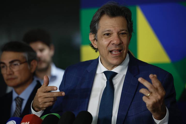 Um homem de terno azul claro está falando em uma coletiva de imprensa， gesticulando com as mãos. Ao fundo， há um painel colorido com as cores verde e amarelo. Outro homem， com cabelo grisalho e óculos， está ao lado， observando.