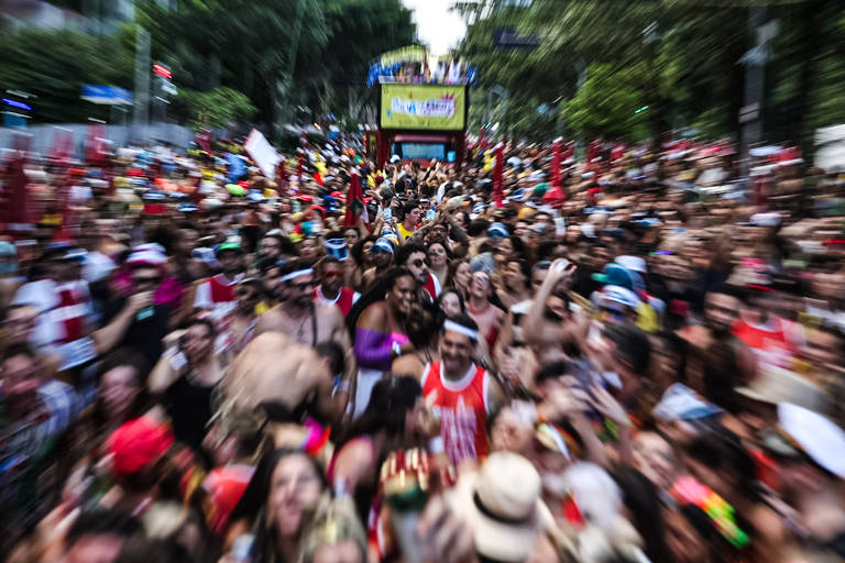 A imagem mostra uma grande multidão durante uma celebração de carnaval, com pessoas vestindo roupas coloridas e algumas sem camisa. Ao fundo, há um carro de som decorado, e a cena é cheia de energia e movimento, com bandeiras e adereços típicos do carnaval. A imagem está levemente desfocada, transmitindo a sensação de agitação e festa.