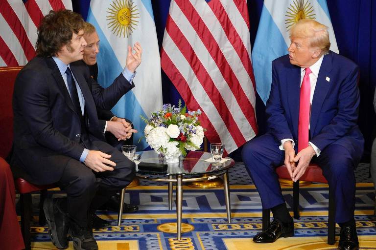 A imagem mostra uma reunião entre três homens em um ambiente formal. Dois homens estão sentados em cadeiras， enquanto um deles gesticula com a mão. Ao fundo， há bandeiras da Argentina e dos Estados Unidos. Uma mesa de vidro com um arranjo de flores está entre eles.