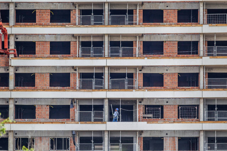 A imagem mostra um edifício em construção com várias andares. As paredes são feitas de tijolos expostos e há janelas com grades de proteção. Um trabalhador está visível em um dos andares， utilizando um equipamento de pintura. O cenário é urbano， com a estrutura ainda em fase de finalização.