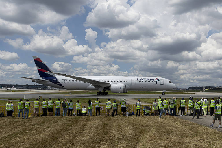 A imagem mostra um avião da LATAM， modelo Boeing 787， taxiando na pista de um aeroporto. Em primeiro plano， há um grupo de pessoas， possivelmente entusiastas da aviação， usando coletes amarelos e observando o avião. O céu está parcialmente nublado， com nuvens brancas e um fundo azul. A grama está visível na parte inferior da imagem.