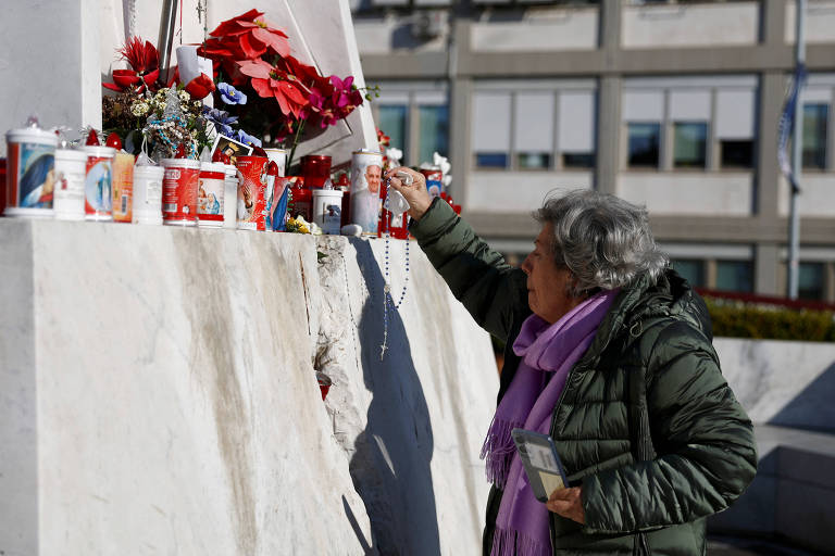 Uma mulher idosa， vestindo um casaco verde e um cachecol roxo， está acendendo uma vela em um memorial. O memorial é decorado com flores e velas， e ao fundo há um edifício com janelas. A mulher parece concentrada em sua ação de homenagem.