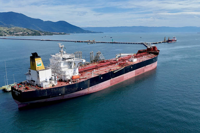 A imagem mostra um grande navio tanque ancorado em águas calmas， com um porto ao fundo. O navio possui um casco preto e uma superestrutura branca， com detalhes em amarelo. Montanhas verdes são visíveis ao fundo， e há outros barcos e estruturas portuárias na área.