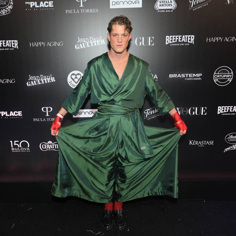 A imagem mostra uma pessoa em um evento de moda, vestindo um robe verde escuro com mangas largas e uma faixa na cintura. A pessoa está em uma pose que destaca o robe, com as mãos abertas e usando luvas vermelhas. O fundo é uma parede preta com logotipos de várias marcas, incluindo Vogue e Beefeater.