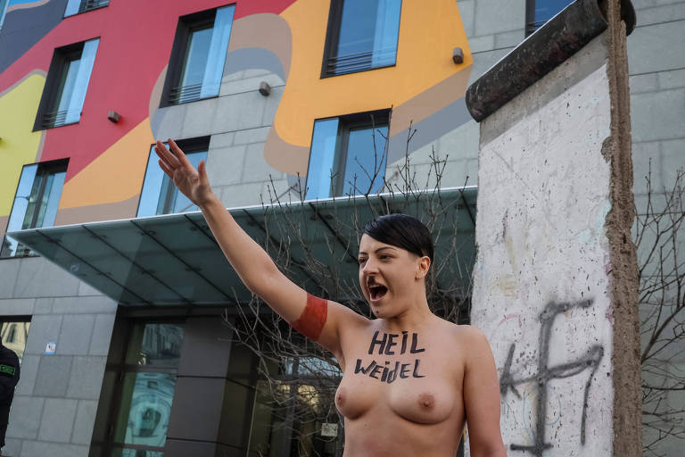 A imagem mostra uma mulher nua em pé em frente a um edifício com um mural colorido. Ela está levantando o braço direito em um gesto de saudação, enquanto grita. Na sua pele, está escrito 'HEIL WEIDEL'. Ao lado dela, há uma parte de um muro com grafite.