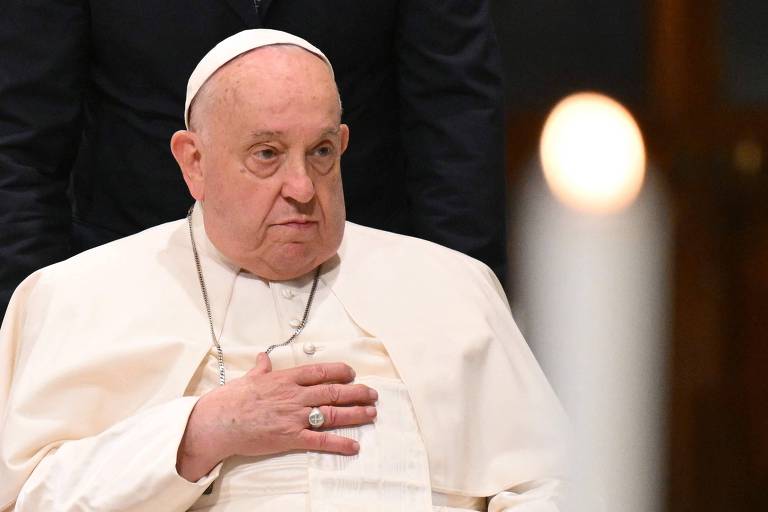 Imagem do Papa Francisco， vestido com vestes brancas e um manto， sentado e com a mão sobre o peito. Ele parece estar em um ambiente solene， com uma vela acesa visível ao fundo. A expressão facial do Papa é séria e contemplativa.