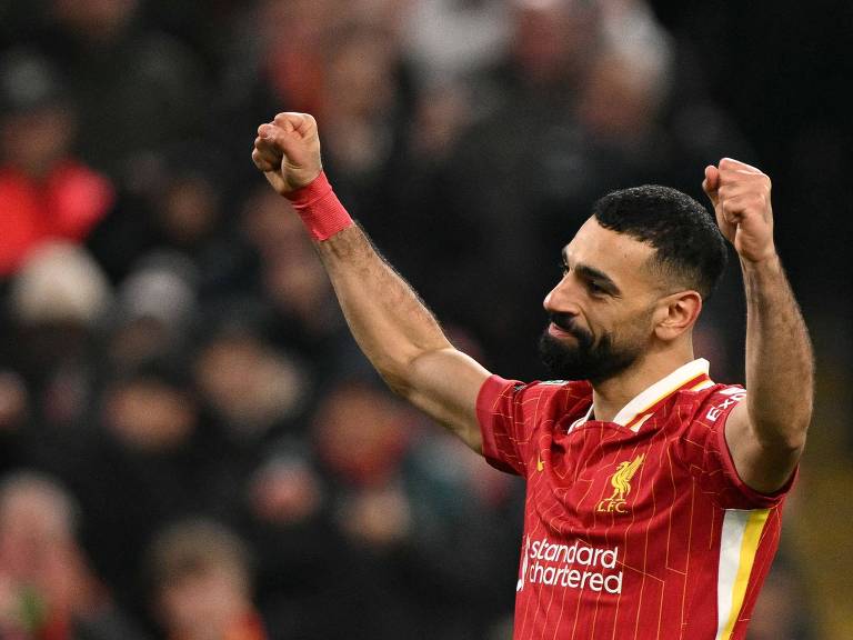 O jogador de futebol Mohamed Salah， do Liverpool， está comemorando com os braços levantados. Ele veste uma camisa vermelha com detalhes em branco e um emblema no peito. O fundo mostra uma multidão de torcedores， com algumas pessoas visíveis em um estádio.