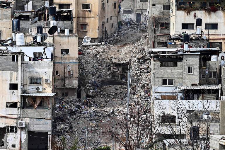 A imagem mostra uma área urbana devastada， com prédios danificados e em ruínas. Há escombros visíveis no centro da cena， onde parece ter ocorrido um deslizamento ou demolição. Os edifícios ao redor apresentam janelas quebradas e estruturas comprometidas， com alguns telhados danificados. A cena transmite um forte sentimento de destruição e abandono.