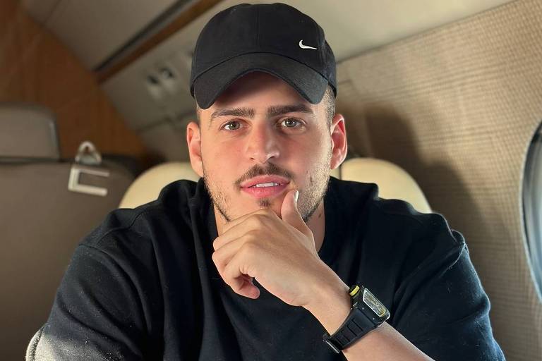  Um jovem sentado em um assento de um jato particular. Ele está usando uma camiseta preta com texto em branco e um boné preto. O jovem tem uma expressão pensativa, com a mão apoiada no queixo. Ao fundo, é possível ver a janela do jato e parte do interior, que é revestido em tons claros e madeira.