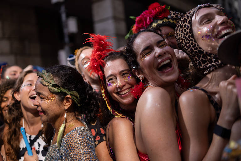 A imagem mostra um grupo de pessoas se divertindo em um ambiente festivo, provavelmente durante o Carnaval. Elas estão sorrindo e se abraçando, com algumas usando adereços coloridos, como penas e glitter. O fundo é desfocado, mas sugere uma atmosfera animada e alegre.