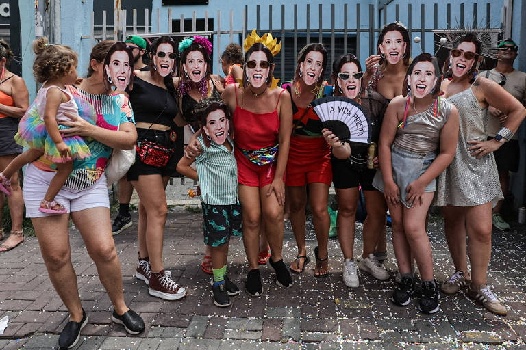 Um grupo de mulheres e crianças posando para a foto em um ambiente festivo. Todas estão usando máscaras com rostos de uma mesma pessoa， além de roupas coloridas e acessórios de carnaval. Algumas mulheres seguram leques e há confete no chão. Uma criança está sendo segurada por uma mulher à esquerda.