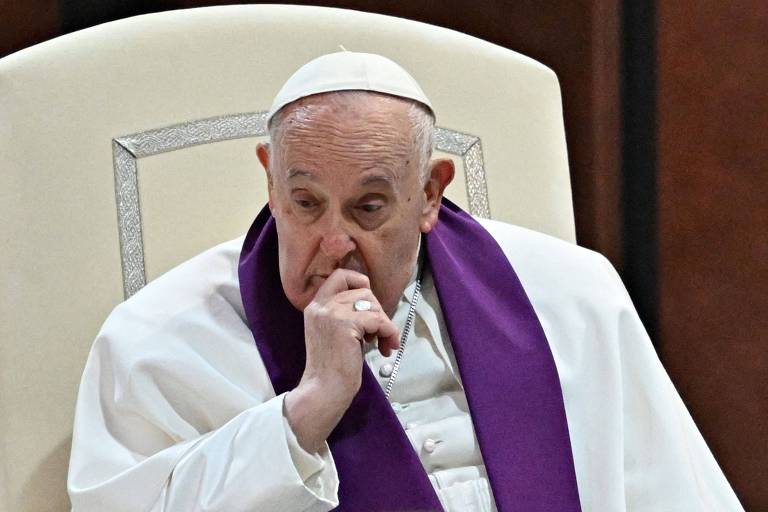 O Papa Francisco está sentado em uma cadeira， vestindo uma túnica branca e um manto roxo. Ele está com a mão no rosto， parecendo pensativo. O fundo é de um ambiente interno， com paredes de madeira.