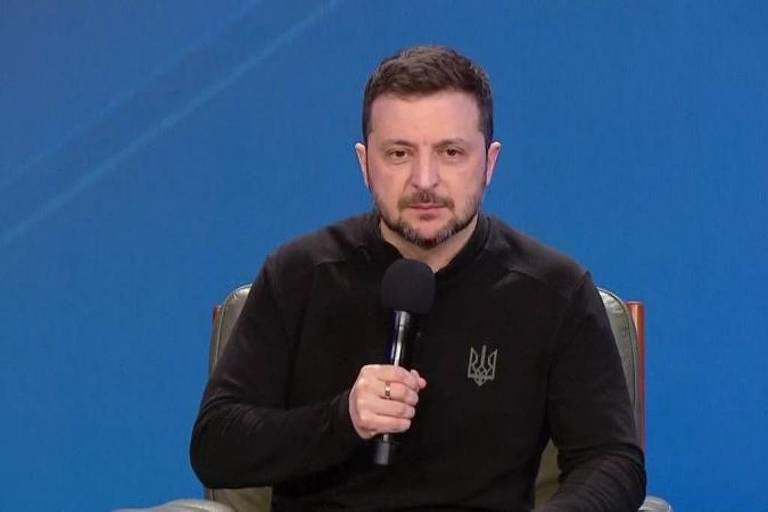 Um homem sentado em uma cadeira， segurando um microfone. Ele está usando uma camiseta preta com um símbolo em destaque. O fundo é azul， e ele parece estar em um ambiente de entrevista ou conferência.