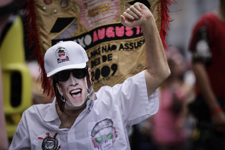 Um homem está levantando o braço em um gesto de celebração ou protesto. Ele usa uma camisa branca com detalhes e uma máscara que representa um rosto sorridente. O homem também está usando um boné branco e óculos escuros. Ao fundo， há uma bandeira ou cartaz com texto visível， mas não legível.