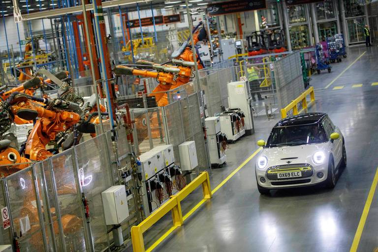 A imagem mostra um ambiente de fábrica de automóveis， com um carro pequeno e moderno， de cor preta e detalhes em amarelo， circulando por um piso de concreto. Ao fundo， há robôs industriais laranja em uma linha de montagem， cercados por painéis de proteção. O ambiente é bem iluminado e organizado， com equipamentos e ferramentas visíveis ao redor.