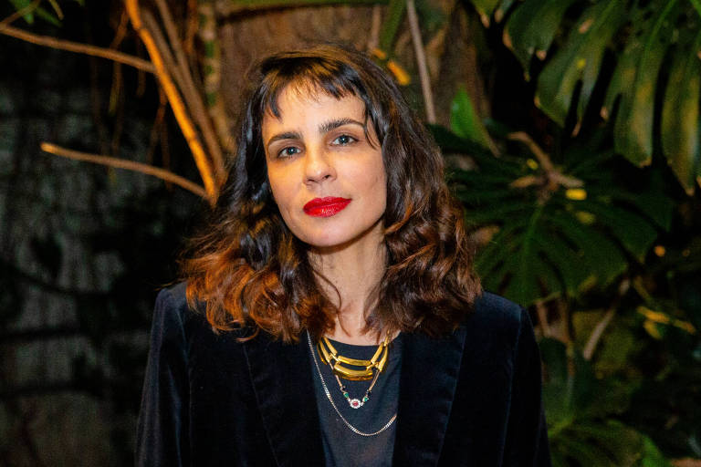 Uma mulher com cabelo castanho escuro e ondulado, usando um blazer preto e uma blusa preta, está em um ambiente com plantas verdes ao fundo. Ela tem lábios vermelhos e está olhando para a câmera.