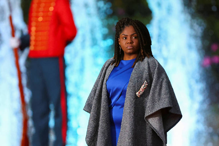 Uma mulher de pele escura está em pé em um tapete vermelho， vestindo um vestido azul e uma capa cinza. Ao fundo， há uma fonte com água jorrando e um soldado em uniforme vermelho e azul， segurando uma arma. A cena é iluminada e festiva.