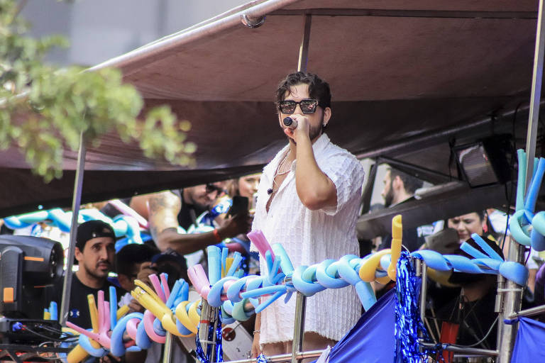 Um artista está se apresentando em um palco ao ar livre， segurando um microfone. Ele usa óculos escuros e uma camisa branca. Ao fundo， há várias pessoas assistindo e algumas estão interagindo. O palco está decorado com fitas coloridas e balões. A atmosfera parece festiva.