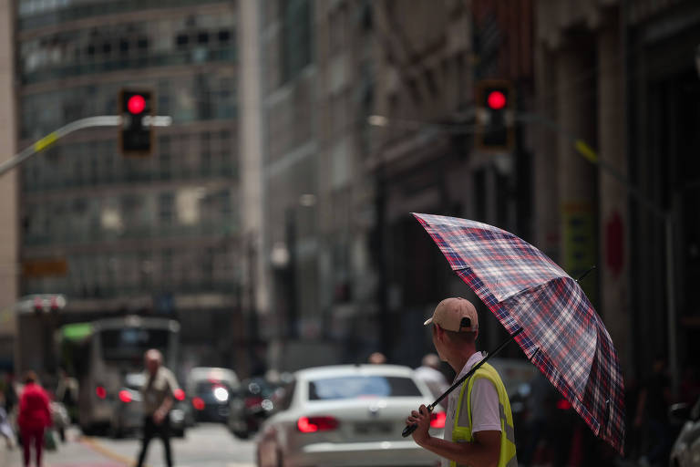 A imagem mostra um homem com um guarda-chuva xadrez em um cruzamento urbano. Ele está de pé na calçada， observando o tráfego. Ao fundo， há veículos em movimento e semáforos vermelhos. O ambiente é urbano， com prédios altos e uma iluminação intensa， sugerindo um dia ensolarado.