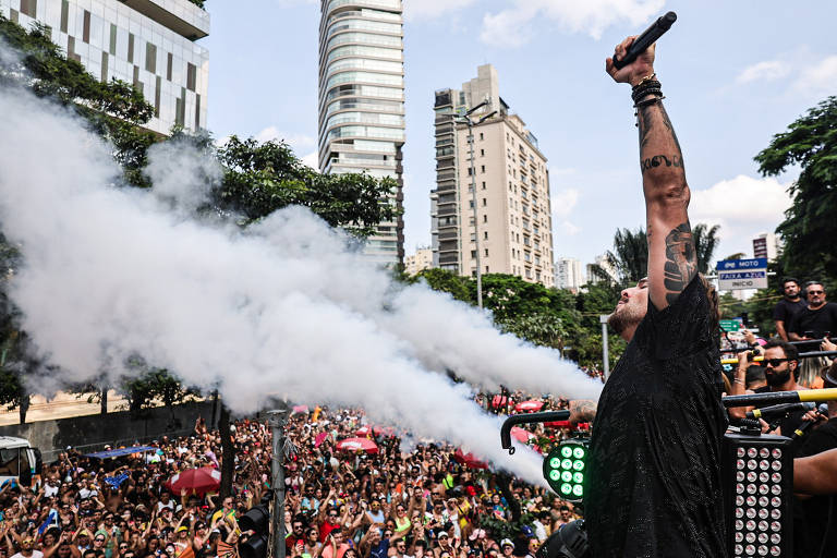 A imagem mostra um artista em um palco, levantando um microfone com uma mão, enquanto fumaça branca é expelida de máquinas ao seu redor. Abaixo, uma grande multidão de pessoas se reúne, algumas segurando guarda-chuvas coloridos. Ao fundo, há prédios altos e vegetação.