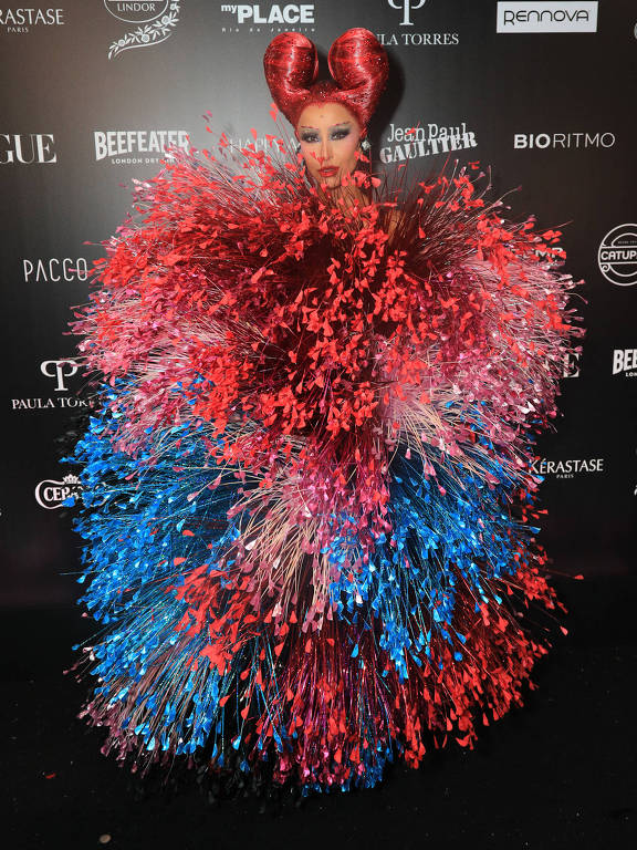 A imagem mostra uma pessoa em um vestido volumoso e colorido, composto por uma mistura de cores vermelha, azul e rosa. O vestido é feito de elementos que se assemelham a penas ou fitas, criando um efeito de explosão. A pessoa tem um penteado elaborado, com cabelo vermelho em formato de coração. O fundo é escuro, com logotipos de marcas de moda visíveis.