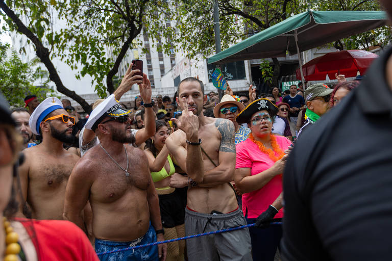 A imagem mostra uma multidão reunida em um evento ao ar livre. Algumas pessoas estão sem camisa, enquanto outras usam roupas coloridas e acessórios. Há uma variedade de expressões faciais entre os participantes, que parecem estar se divertindo. Ao fundo, pode-se ver árvores e barracas, sugerindo um ambiente festivo.