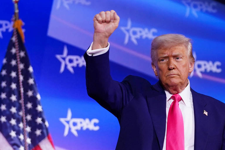 Um homem com cabelo loiro e terno escuro levanta o punho em um palco, enquanto uma bandeira dos Estados Unidos está ao fundo. O fundo é decorado com o logotipo do CPAC em azul e branco.