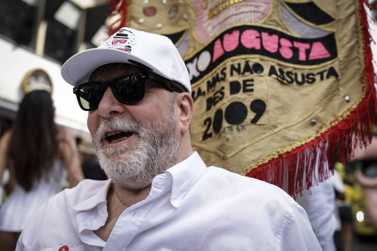 Um homem com barba e cabelo grisalho, usando óculos escuros e um boné branco, sorri enquanto está sentado. Ele veste uma camisa branca e está em um ambiente festivo, com uma bandeira ao fundo que diz 'A AUGUSTA NÃO ASSUSTA DESDE 2009'.